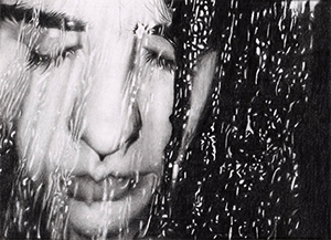a black and white image of a person with their eyes closes, the face is distorted by water over a glass surface in front