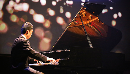 A man plays a piano on a darkened theatre stage, with a speckled visual backdrop