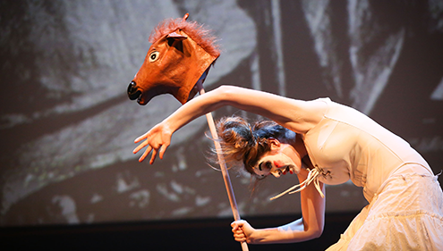 A woman wearing a white dress and white makeup holds a horse head prop with her arms in the air against a grey backdrop