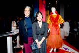 Two women smile in an indoor exhibition space, with a mannequin wearing an orange dress behind them