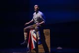 Performer dressed as a First World War private sits on a large wooden crate, which is draped in a Union Jack. Their mouth is open mid-speech.