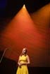 Performer in a canary yellow dress sings a meter away from a standing microphone, lit by a triangle of orange from a spotlight overhead.