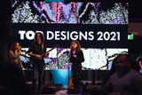 Two singers and a guitarist perform on stage. Behind them is a screen reading 'Top Designs 2021'.