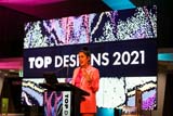 A speaker in a pink jacket stands at a lectern. Behind them is a screen that reads 'Top Designs 2021'.