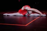 A person with blonde hair tied in a bun wears a red feathery dance costume. She is on the floor in the splits with her head and torso bent backwards while she grabs her back foot with both hands.