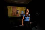 A person with long brown hair wears a blue t-shirt and black pants. She is leaning on a black couch in front of a screen that shows a cartoon of a woman smiling and holding a child wearing an astronaut helmet.