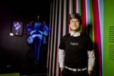 A person with short dark hair with a white streak wears glasses, a black t-shirt that reads 'legacy' over a white collared shirt, black pants, and a white belt. They stand in front of a colourful striped wall next to a display of a black mannequin wearing a blue bodysuit with puffy fabric around the hips and arms.