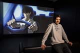 A person with short brown hair wears a grey hoodie and dark jeans. He leans on a black couch in front of a screen that shows someone driving a car in the anime art style.