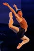 A dancer wears a brown tank top and black shorts. He is in the air mid jump with his arms stretched in a V-shape above his head. His right leg is bent in the front and his left leg is bent back and stretched upward.