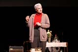 A performer wears a red turtleneck, light brown cardigan, grey pants, and gold jewelry. Her hair and eyebrows are coloured white. She stands by a bar cart and is mid-speech while holding her left hand in front of her torso and her right hand in line with her shoulder.