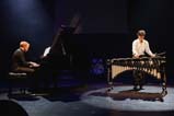 Two musicians are on stage. On the left, a man wearing all black plays the piano. On the right, a performer wears a white shirt and black pants and plays the vibraphone while holding four mallets.