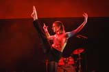 A dancer with slicked back hair wears black pants and a multi-coloured long sleeve shirt. Their arms are outstretched to either side, and their right leg is almost vertical in the air with foot pointed. The stage lights cast a red glow on everything.