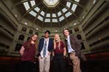 Four Top Talks presenters stand together smiling in the reading room of State Library Victoria. The room has high ceilings with a glass dome at the top. There are three levels of balconies along the walls.