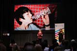 The audience watches a presenter in maroon shirt and black pants on stage with PowerPoint behind them. The PowerPoint shows a boy with dark hair drinking from a plastic water bottle filled with colourful plastic beads.
